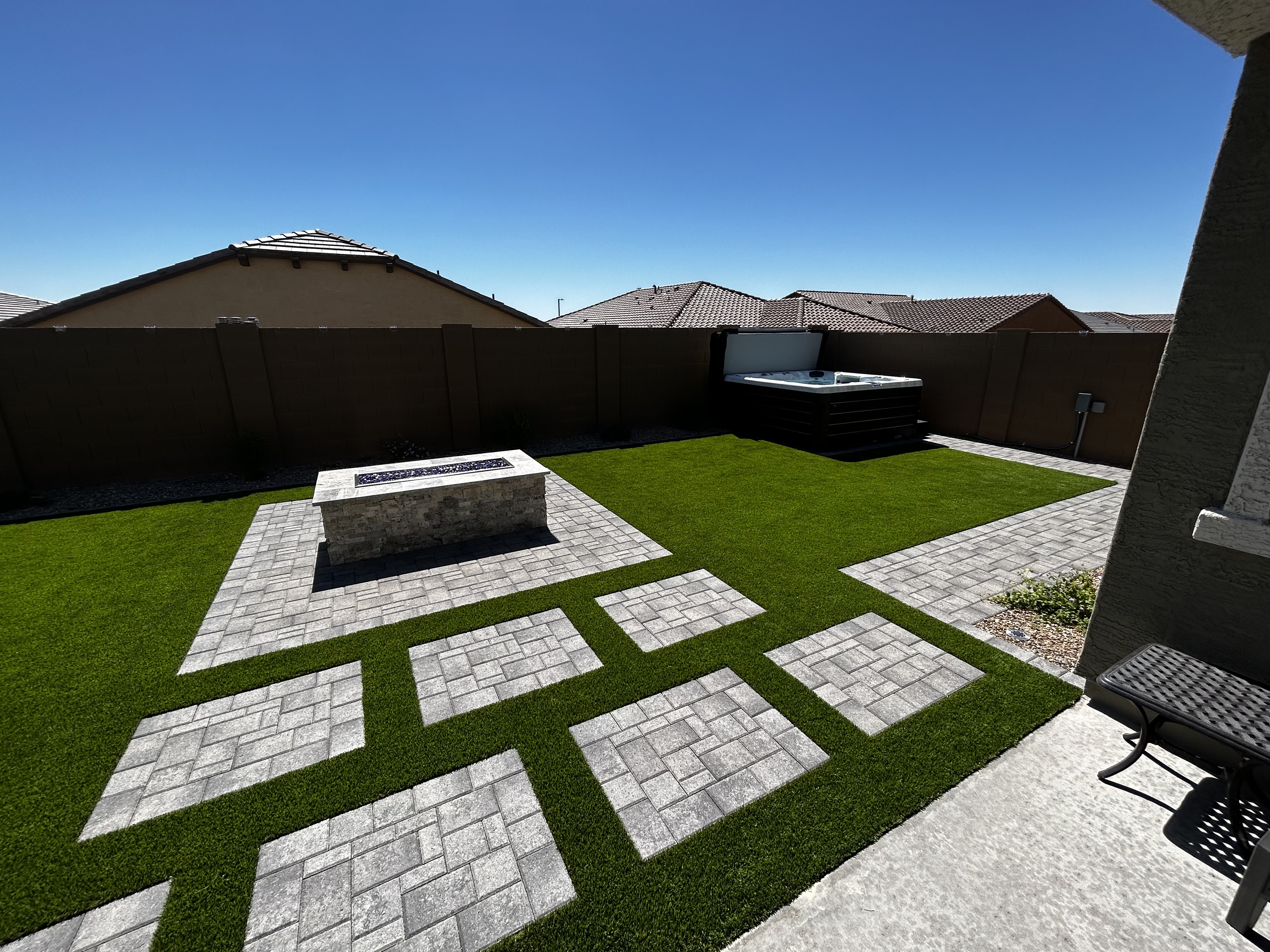Stone fire table on a paver patio with turf inlays and a hot tub against the wall